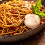 Burst cherry tomato pasta with burrata and basil in a dark skillet
