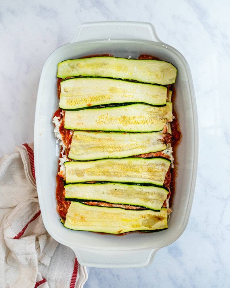 Unbaked lasagna layered with zucchini slices, sauce, and cheese in a white dish